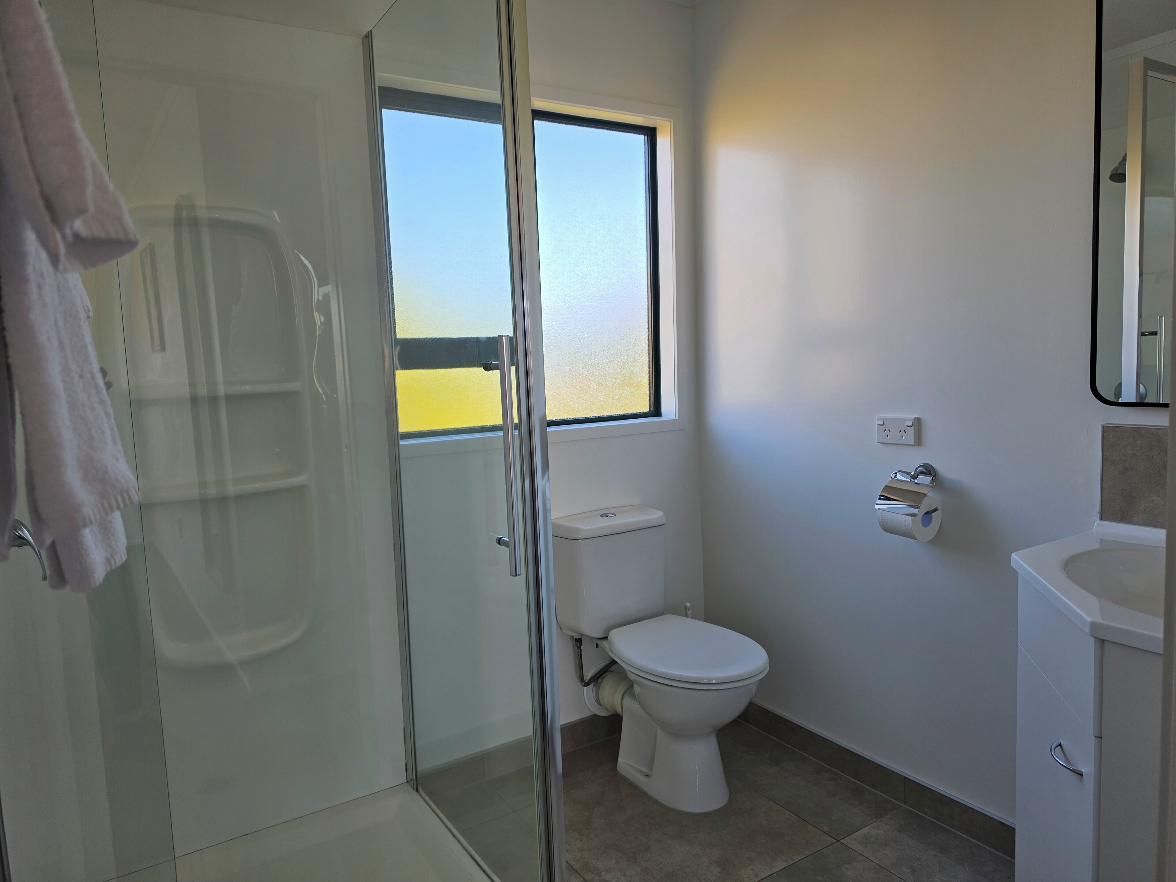 A bathroom with a glass shower enclosure, a toilet, a sink, and a window providing a view.