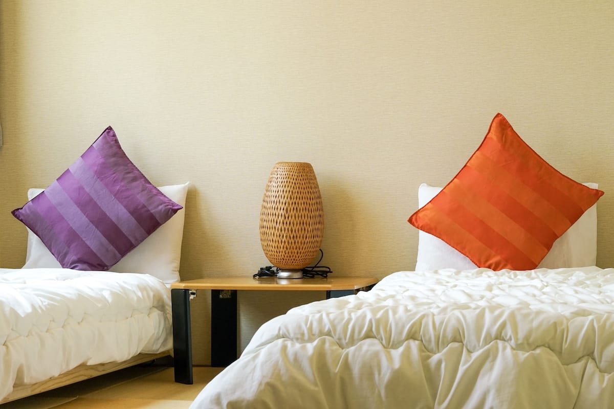 Ein Hotelzimmer mit zwei Betten, jeweils mit weißer Daunendecke, einem kleinen Holztisch mit einer Lampenschirm aus Bändern und zwei Kissen, eines violett und eines orange.