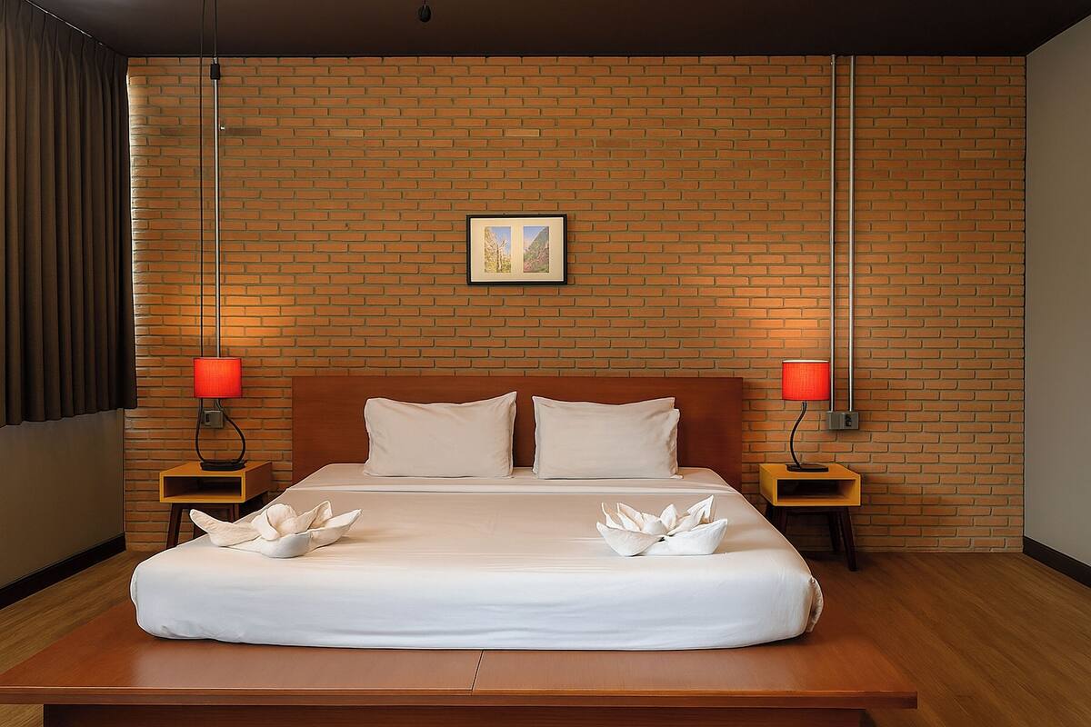 A hotel room with a brick wall, a large bed with white linens, two nightstands with red lamps, and a framed picture on the wall.