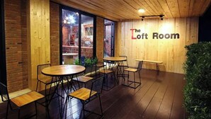 A cozy room with wooden walls and a sign reading "The Loft Room". There are several round tables and chairs arranged for dining or socializing. - The Loft Room Nimman (Chiang Mai)
