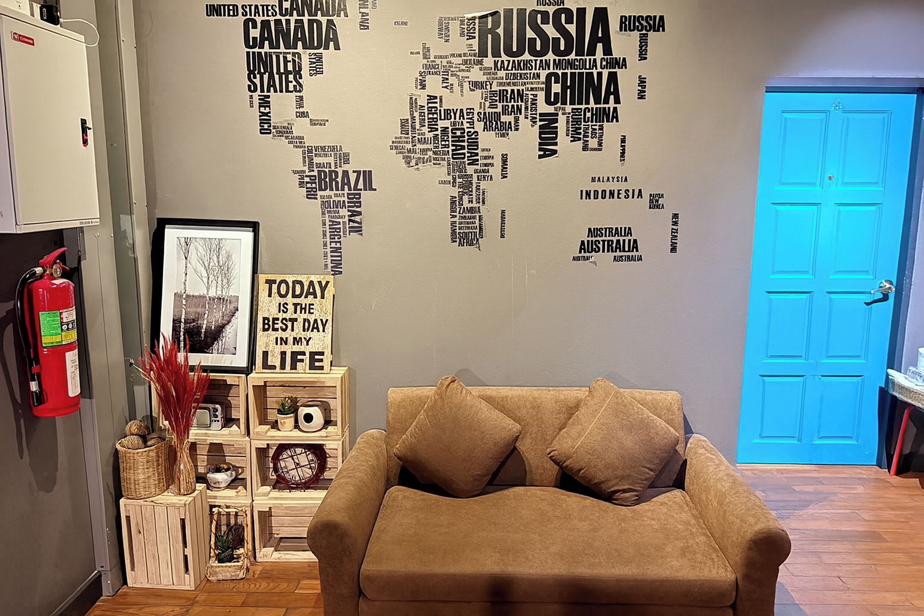 Una habitación con un sofá color beige, un mural de un mapa mundial, un cuadro enmarcado y una puerta azul.