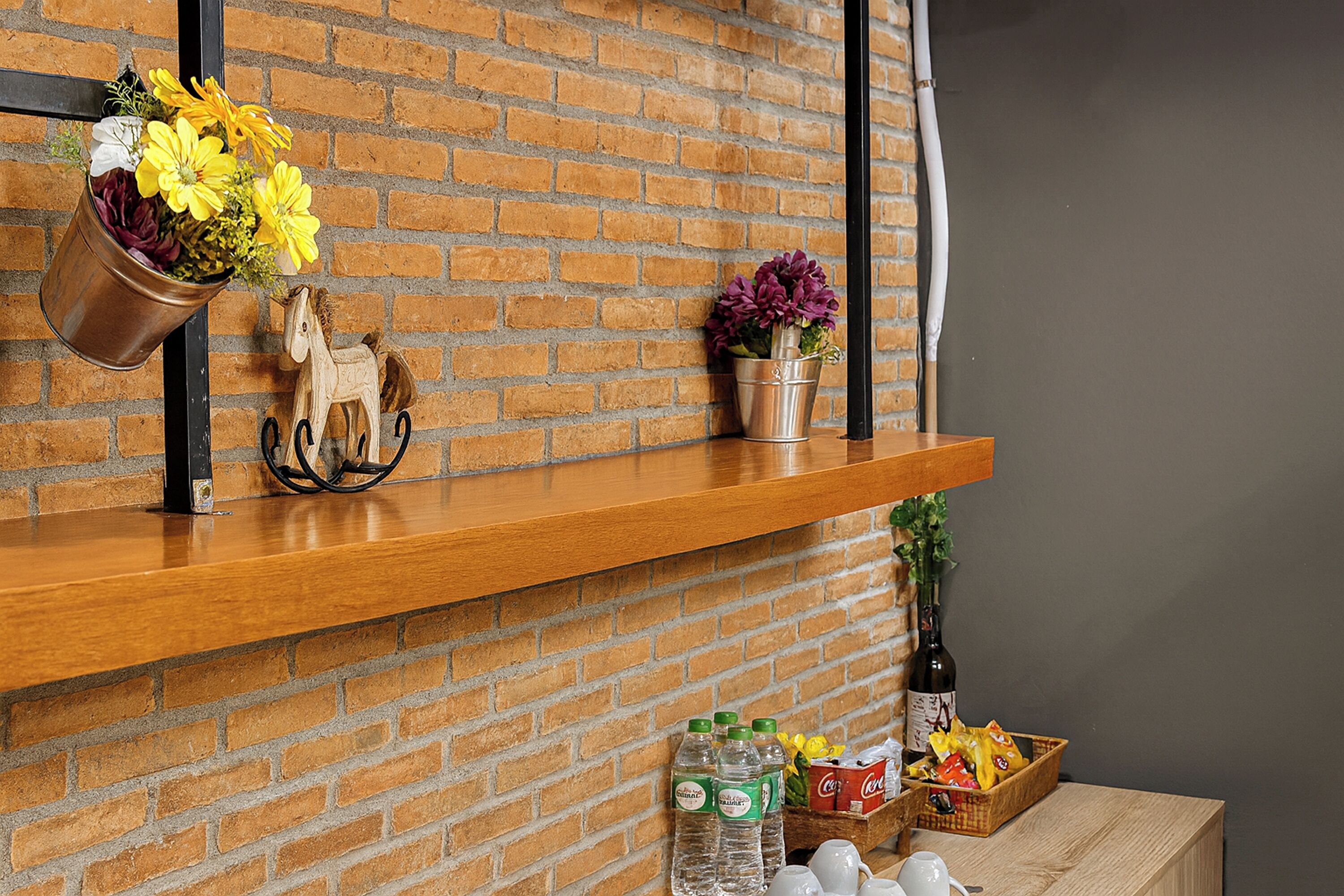 Estante de madera junto a una pared de ladrillo con un cubo metálico lleno de flores, una figurita de caballo y una planta en maceta.