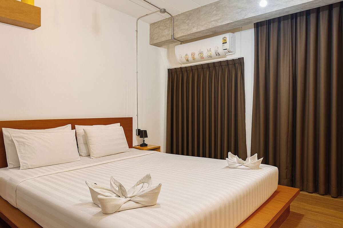 A hotel room with a bed, a nightstand, an air conditioning unit, and brown curtains.
