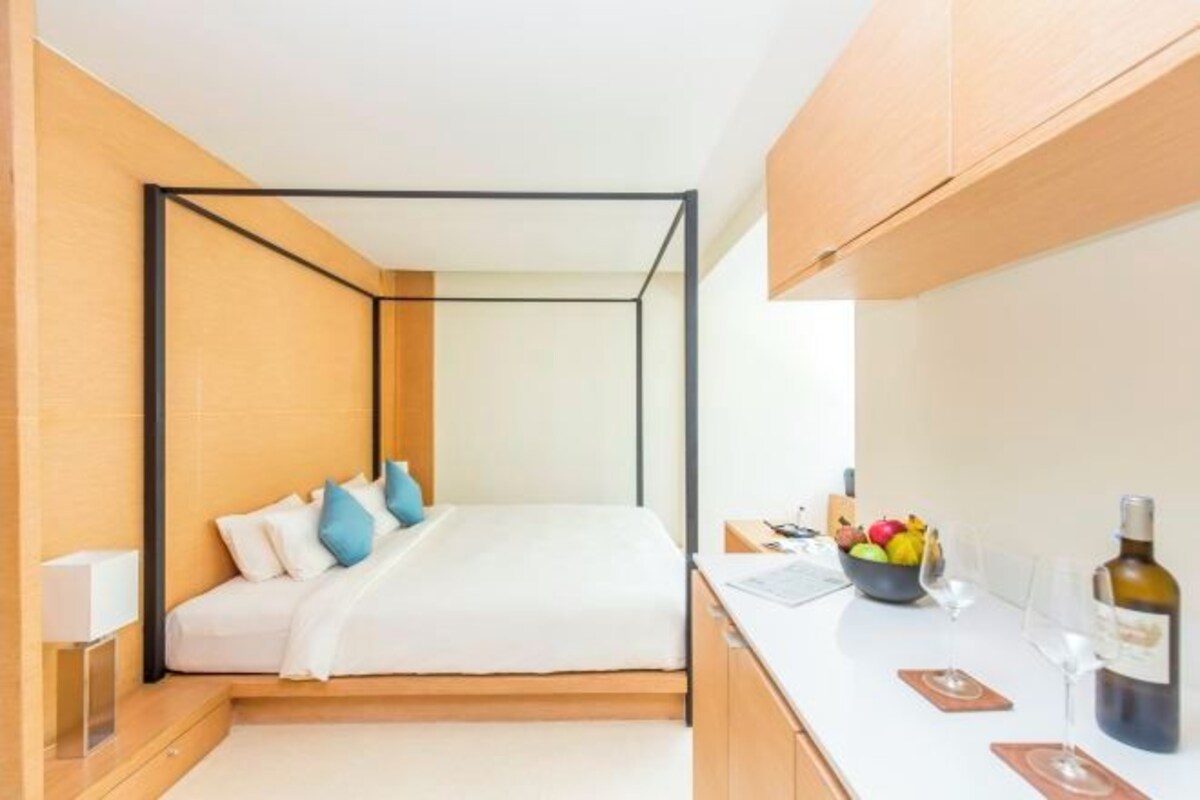 Une chambre moderne au design minimaliste, avec un lit recouvert de draps blancs et agrémenté d’oreillers bleus, une table de chevet sur laquelle se trouve un bol de fruits et une bouteille de vin, et un plan de travail blanc.