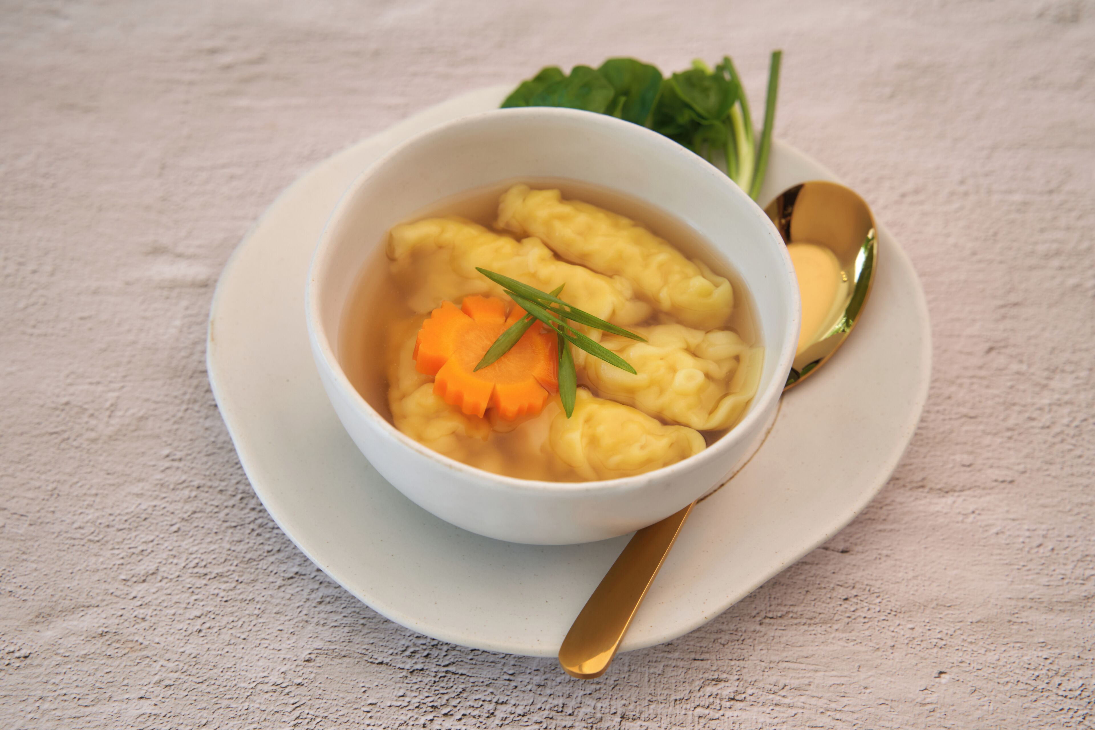Un tazón de sopa con albóndigas y zanahoria, acompañado de una cuchara y un recipiente pequeño de mantequilla sobre un plato blanco.