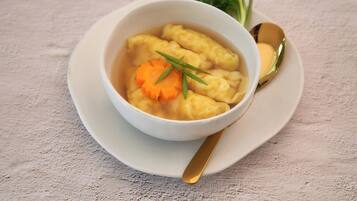 Un tazón de sopa con albóndigas y zanahoria, acompañado de una cuchara y un recipiente pequeño de mantequilla sobre un plato blanco.