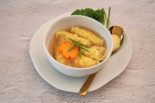 Un tazón de sopa con albóndigas y zanahoria, acompañado de una cuchara y un recipiente pequeño de mantequilla sobre un plato blanco.