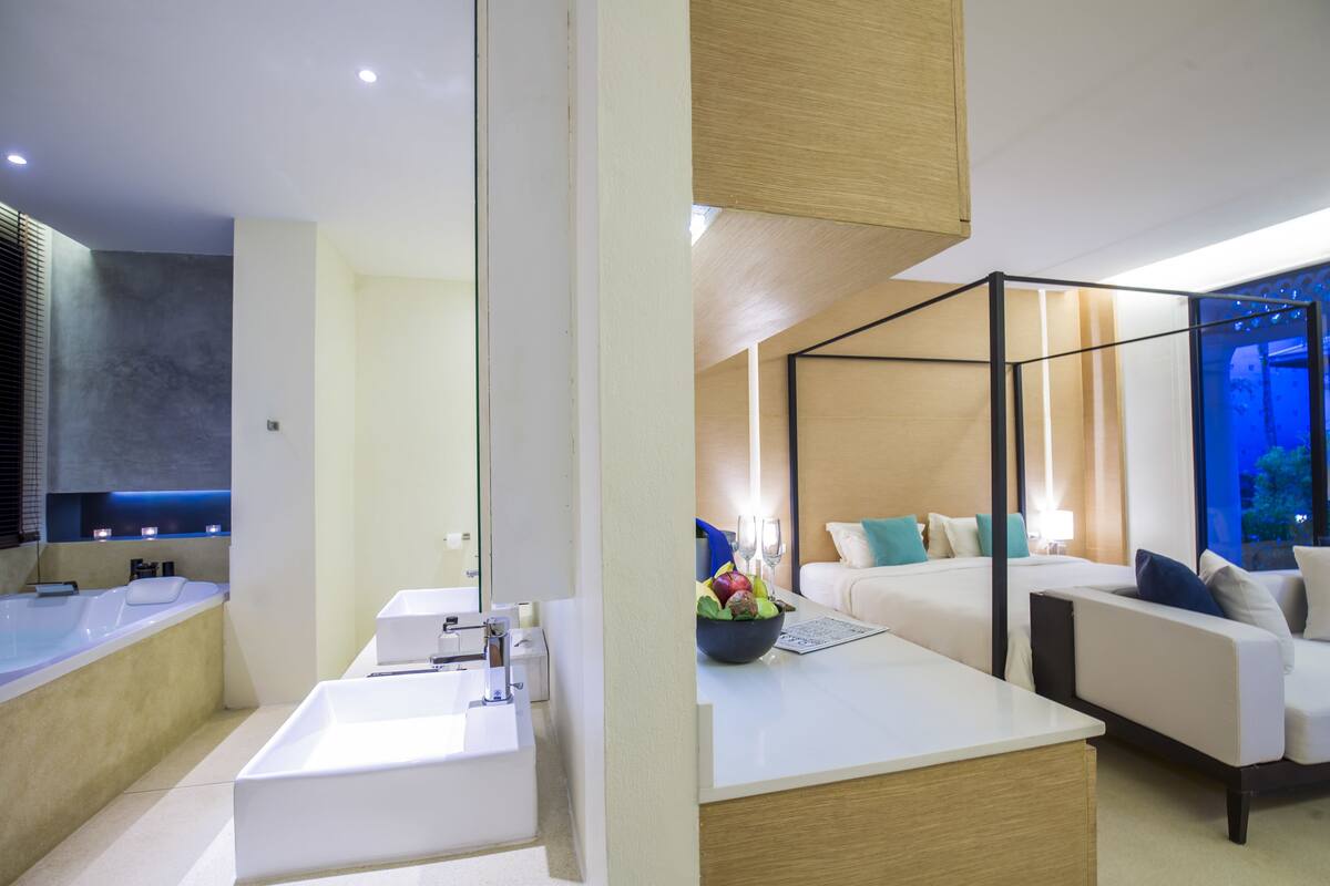 Una habitación de hotel moderna con una bañera grande, zona de ducha separada, espacio de descanso con sofá y mesa de comedor, y un ventanal con vistas al cielo nocturno.