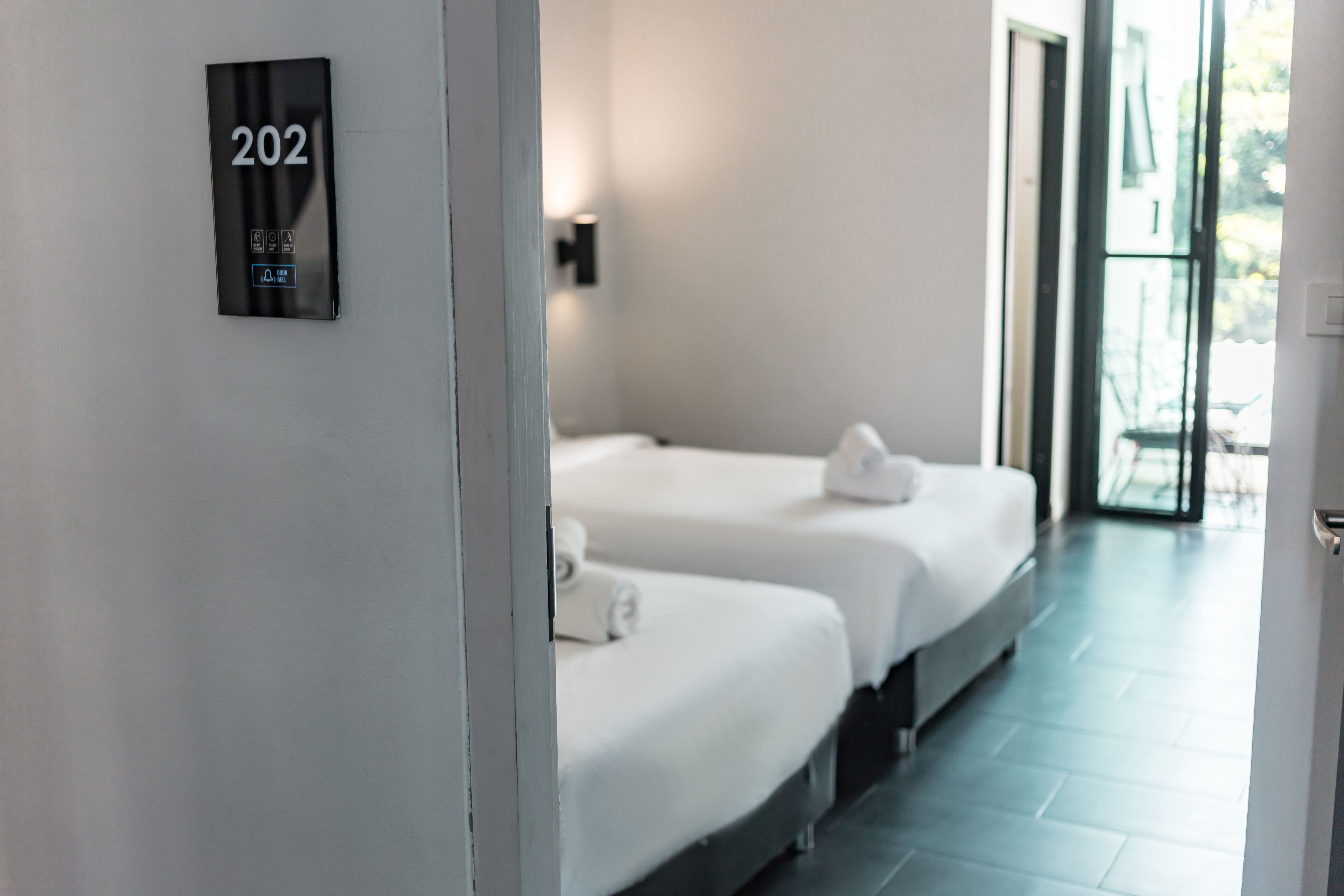 A hotel room with two beds, a number sign '202' on the wall, and a view of the outdoors through a large window.