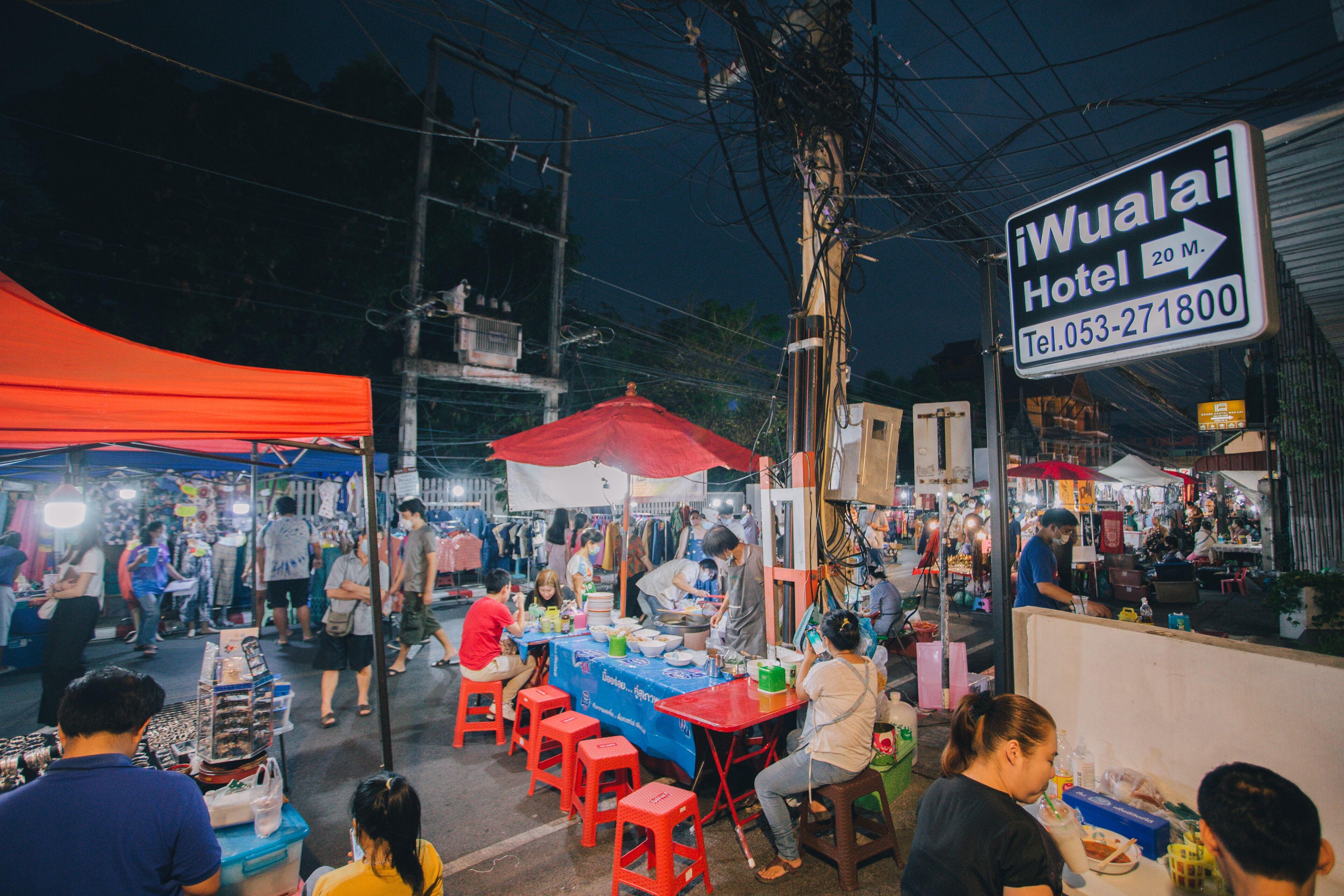 A bustling street market with various stalls and people. A sign for the "iWualai Hotel" is visible, indicating its location.
