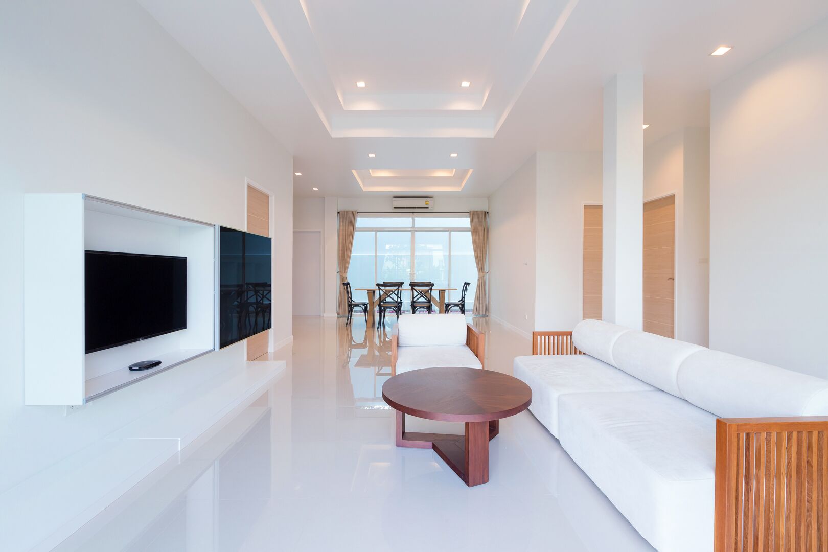 A modern hotel room with a flat-screen TV mounted on the wall, a round wooden coffee table, a white sofa, and a dining area with chairs and a table.