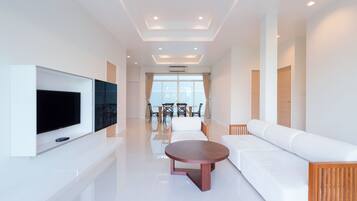 A modern hotel room with a flat-screen TV mounted on the wall, a round wooden coffee table, a white sofa, and a dining area with chairs and a table.