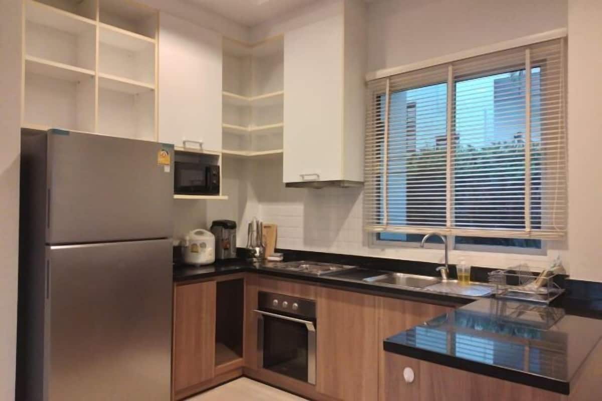 Una cocina moderna con un refrigerador de acero inoxidable, microondas empotrado y horno. Se aprecia una ventana con persianas, un fregadero y una encimera con diversos utensilios de cocina.