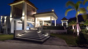 Una casa moderna de dos pisos con una escalera que lleva a la entrada, rodeada de palmeras y un camino de acceso bien iluminado.