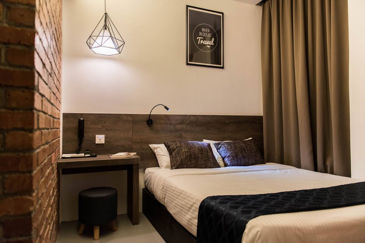 A hotel room with a bed, a desk, a chair, a hanging light fixture, and a framed quote on the wall.