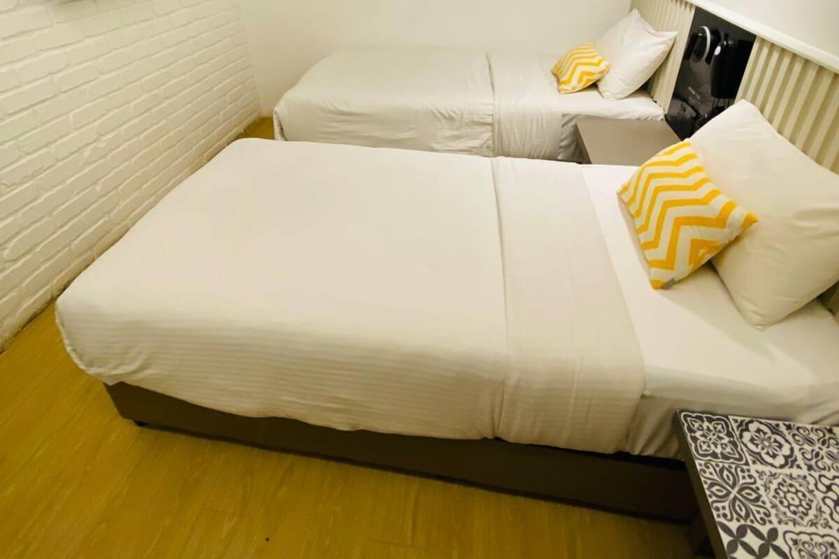 A small, clean room with two single beds, a flat-screen TV mounted on the wall, and a patterned bench.
