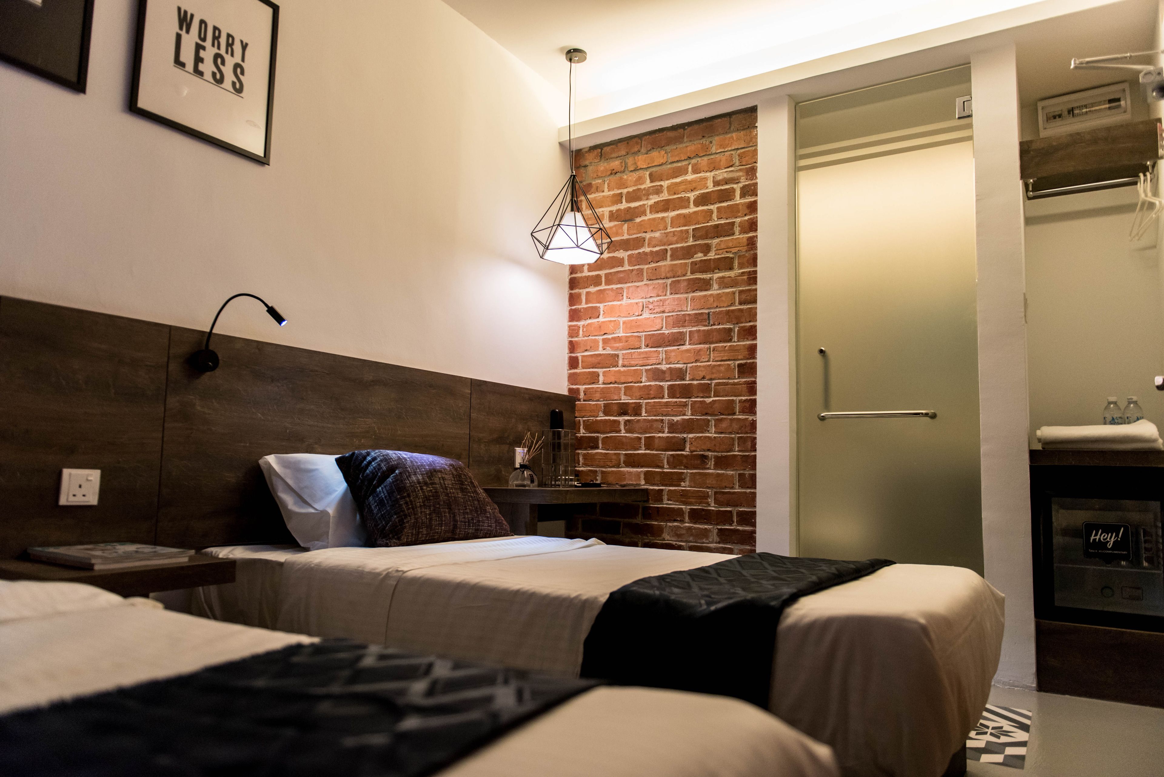 A hotel room with a brick accent wall, a bed with a wooden headboard, a bedside table with a lamp, and a door with a towel rack.