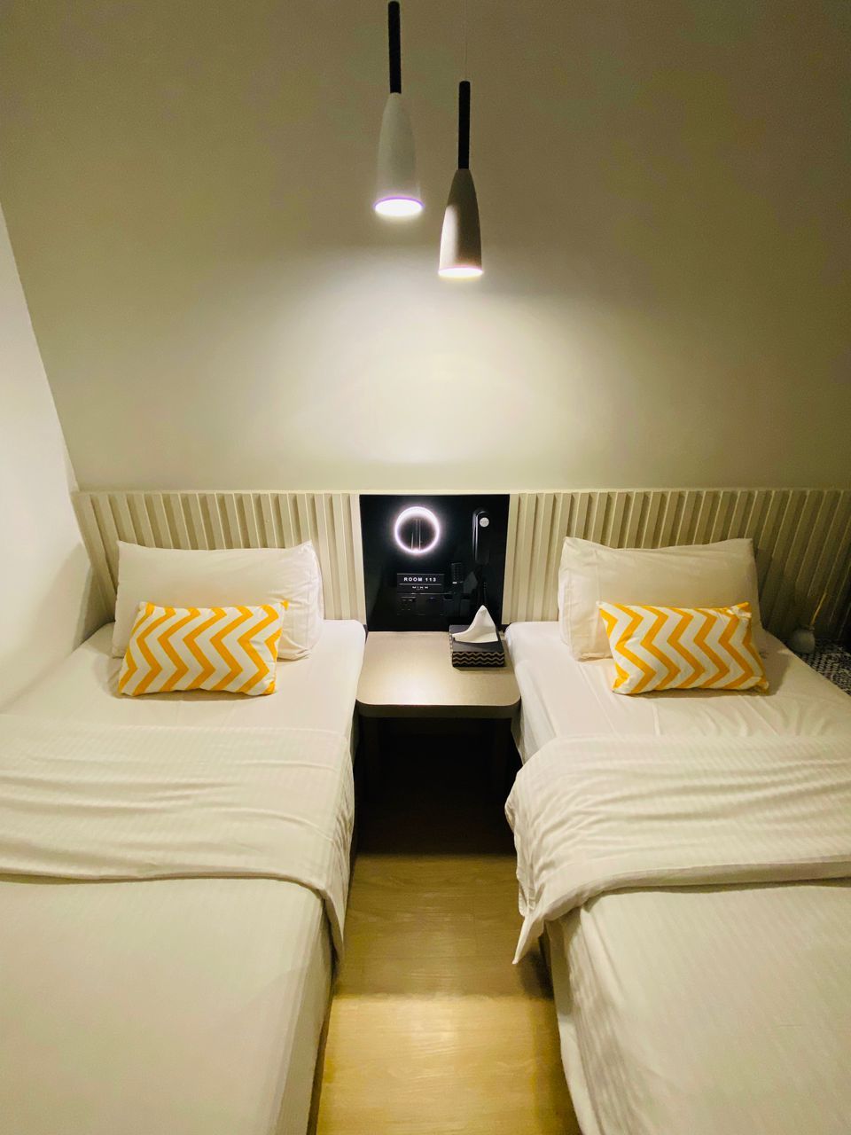 A hotel room with two beds, each with a white bedspread and a yellow and white chevron pillow. There are two pendant lights hanging from the ceiling, and a control panel with a circular light display on the wall between the beds.