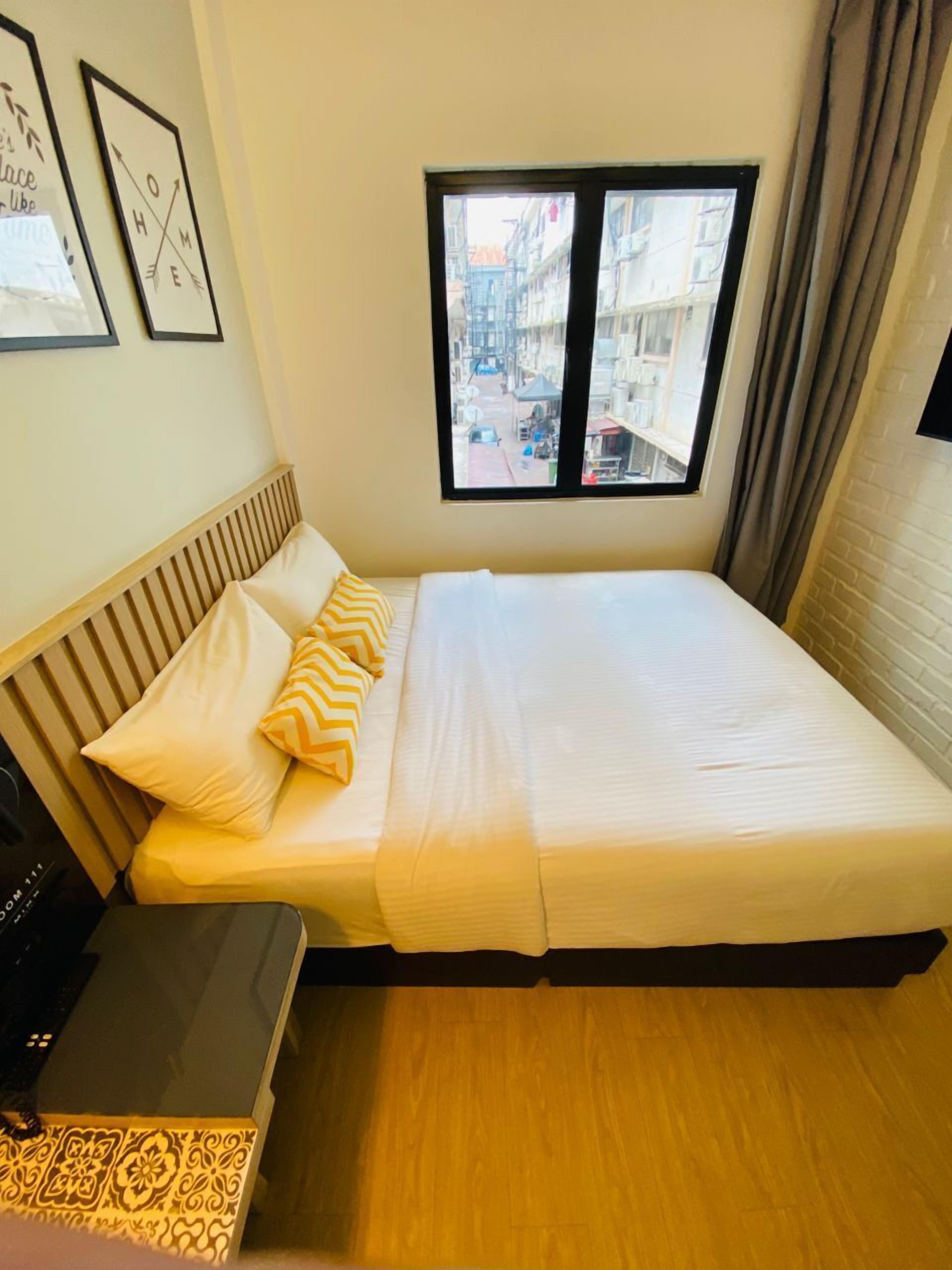 Une chambre à coucher avec un lit, une tête de lit en bois, une fenêtre donnant sur des immeubles et une petite table sur laquelle se trouve une boîte décorative.