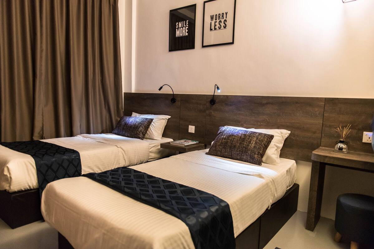 A hotel room with two beds, a nightstand, a desk, and a chair. There are two framed motivational posters on the wall.