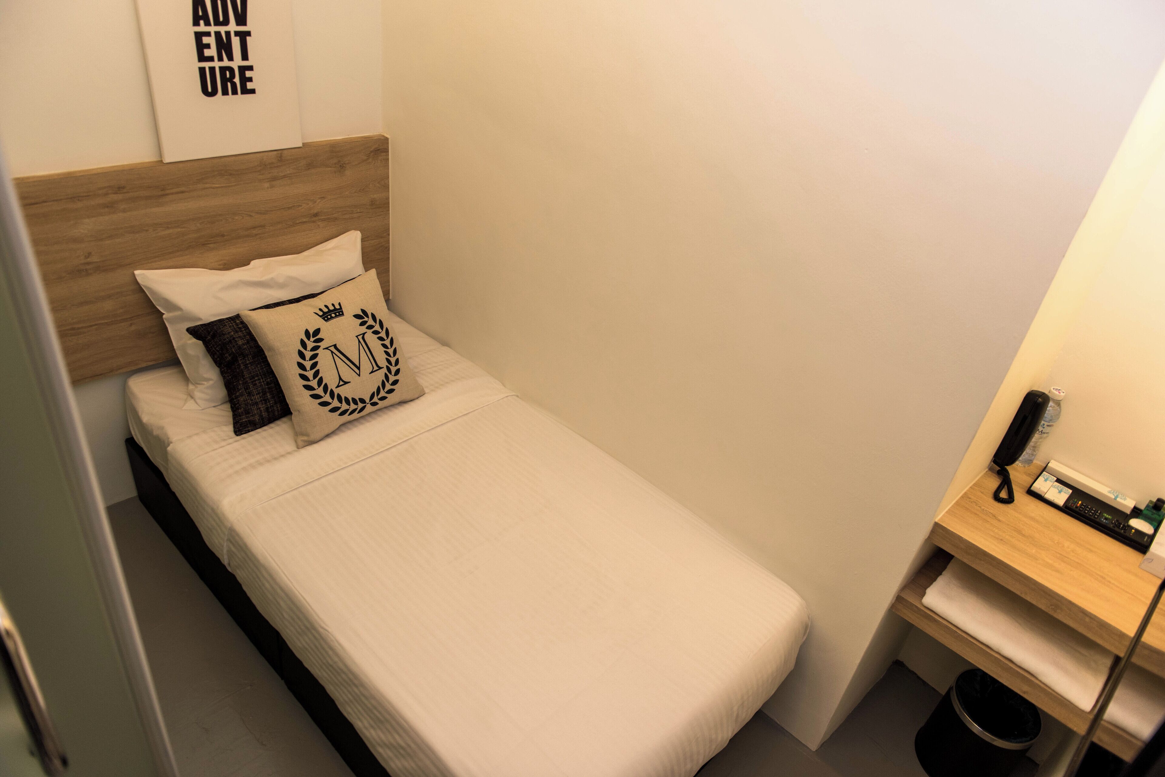 Un petit lit bien fait, avec une tête de lit en bois, une table de chevet sur laquelle se trouvent un téléphone et une petite lampe, et une œuvre d’art accrochée au mur.