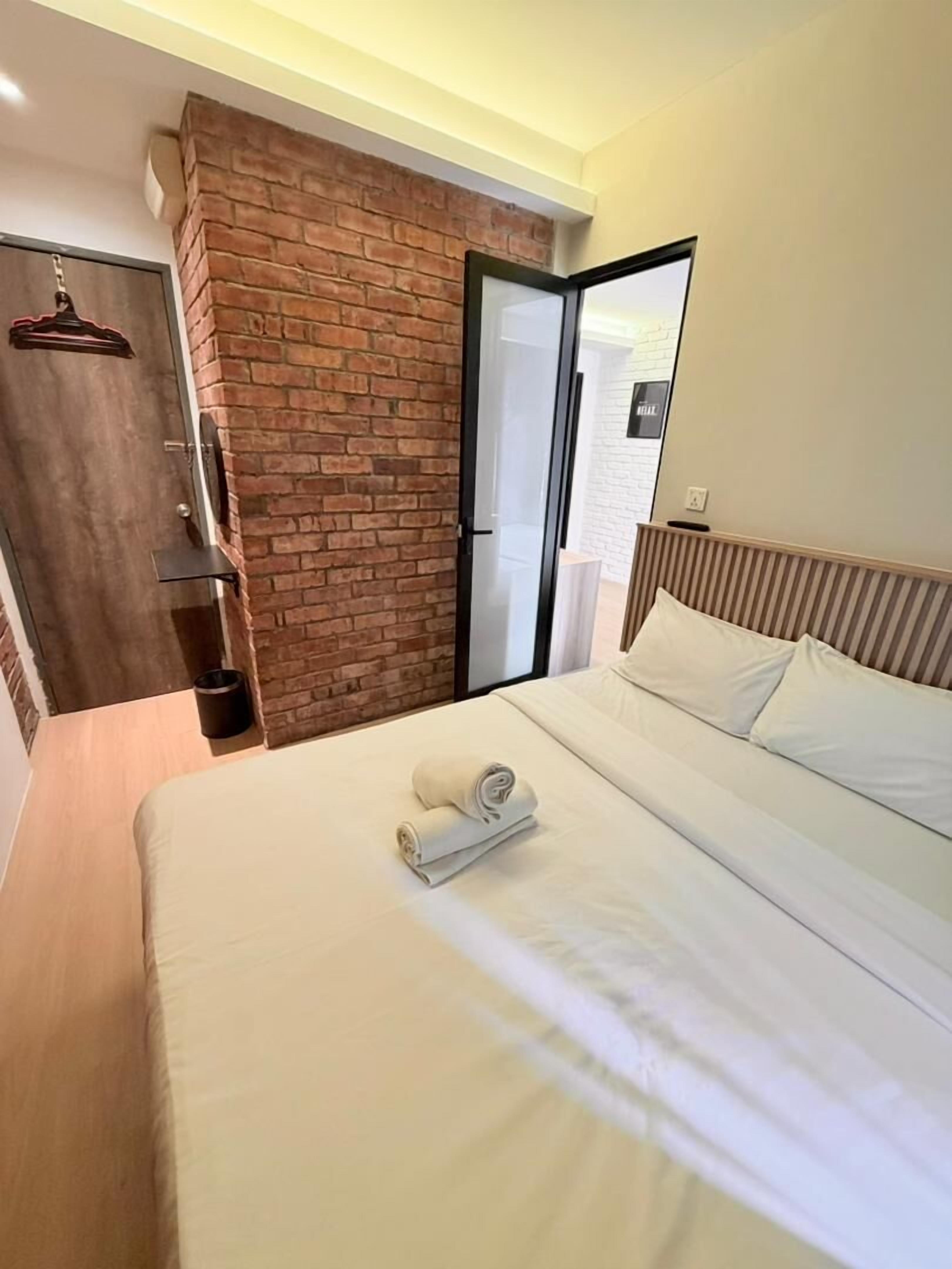 A modern hotel room with a brick accent wall, a large bed with white linens, a wooden headboard, and a sliding door leading to a balcony.