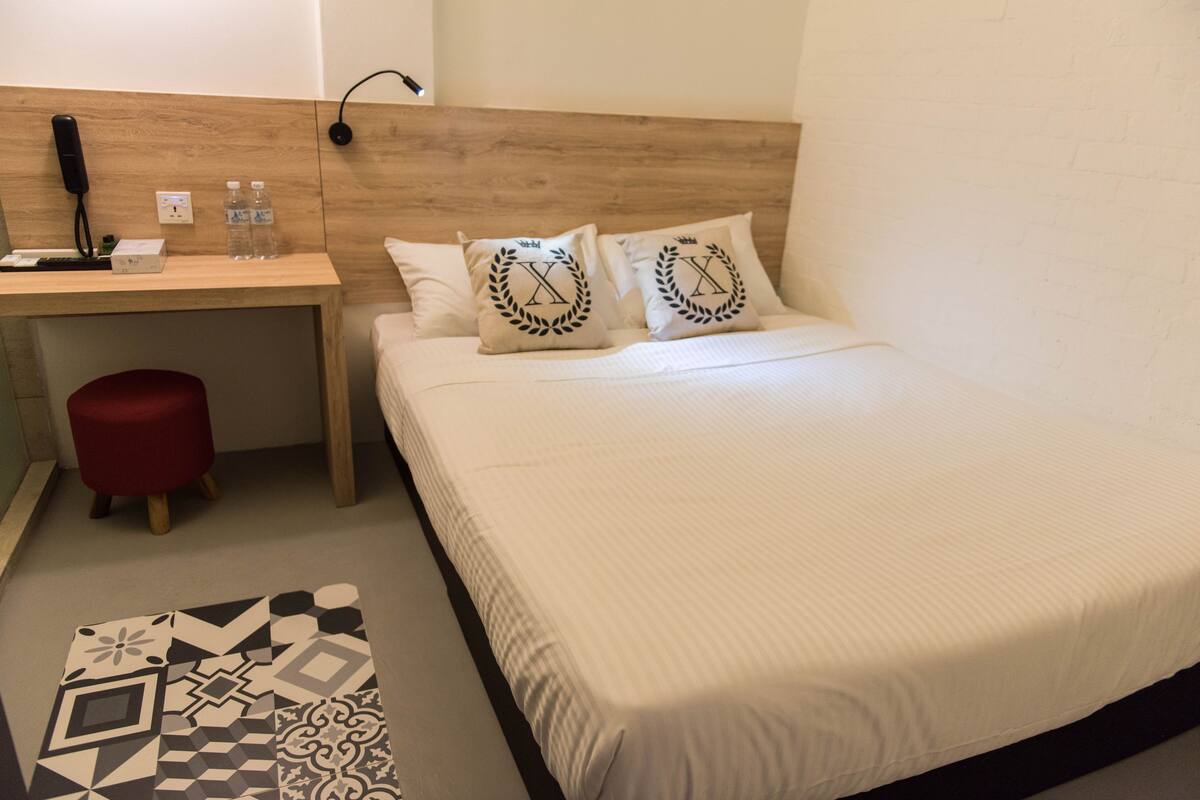 A hotel room with a bed, a desk with a phone, two water bottles, and a red stool.