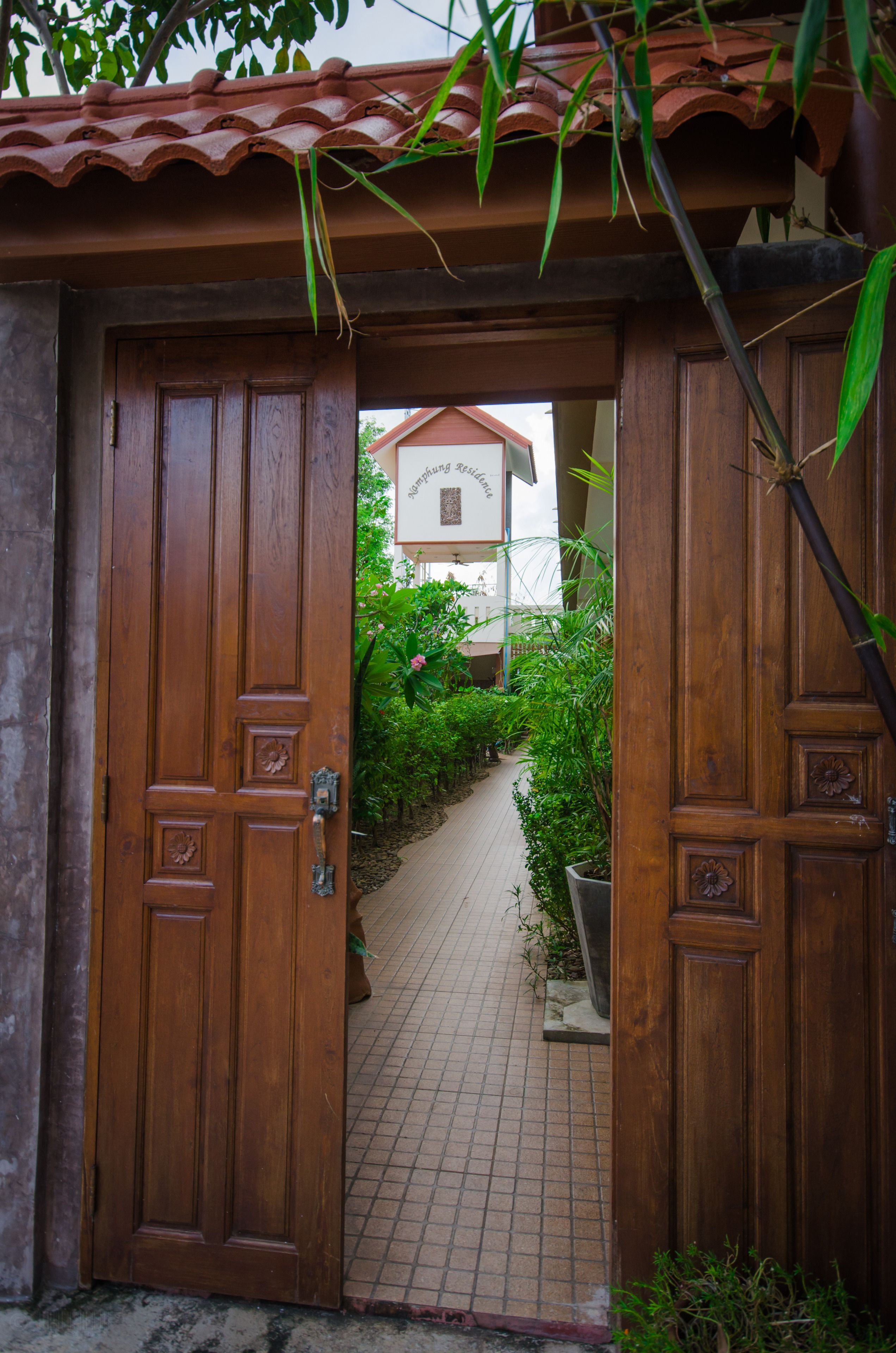 A wooden door leading to a tiled pathway with greenery and a building in the background.