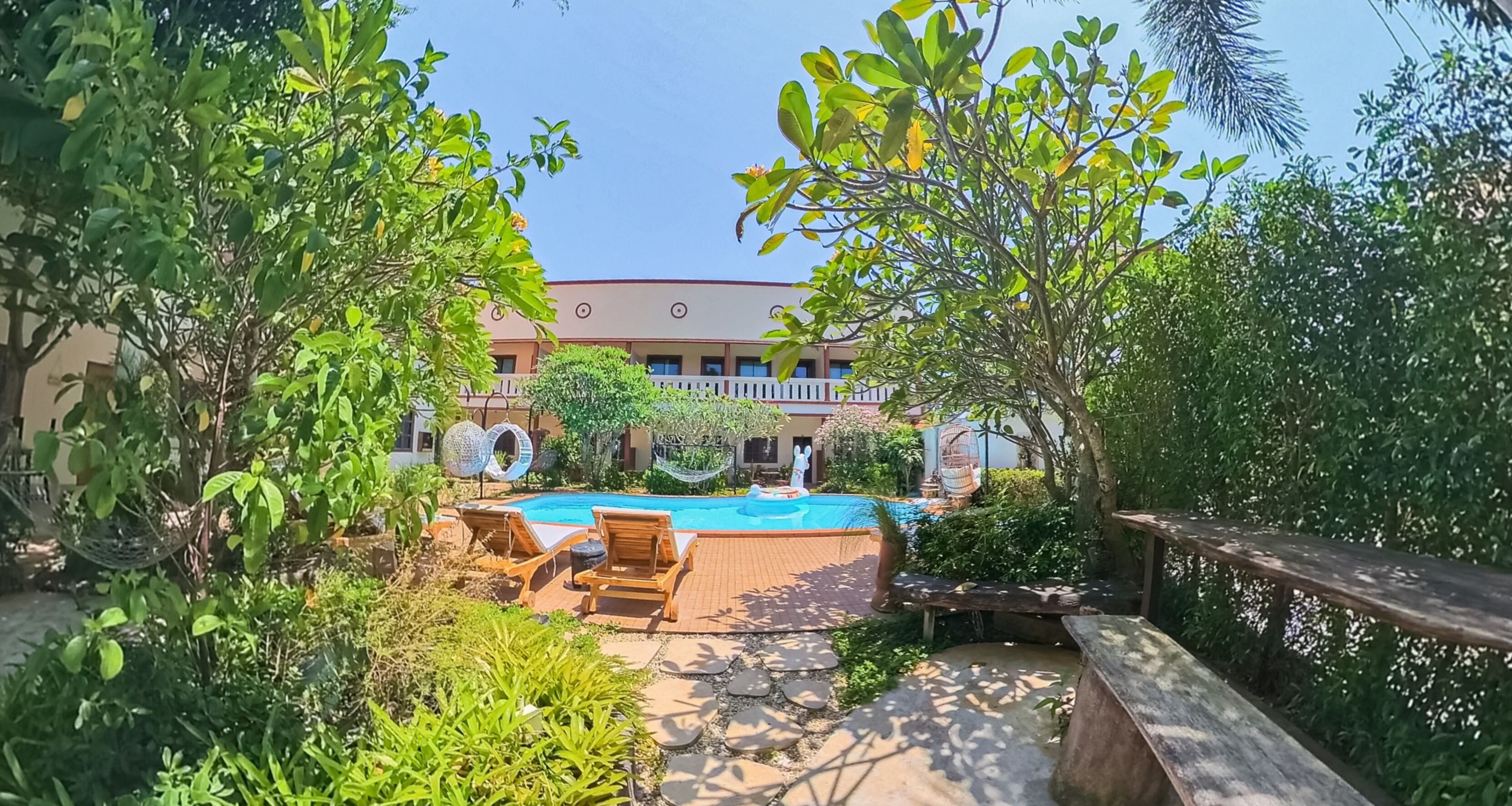 A pool area with lounge chairs, surrounded by lush greenery and a building with a white fence in the background.