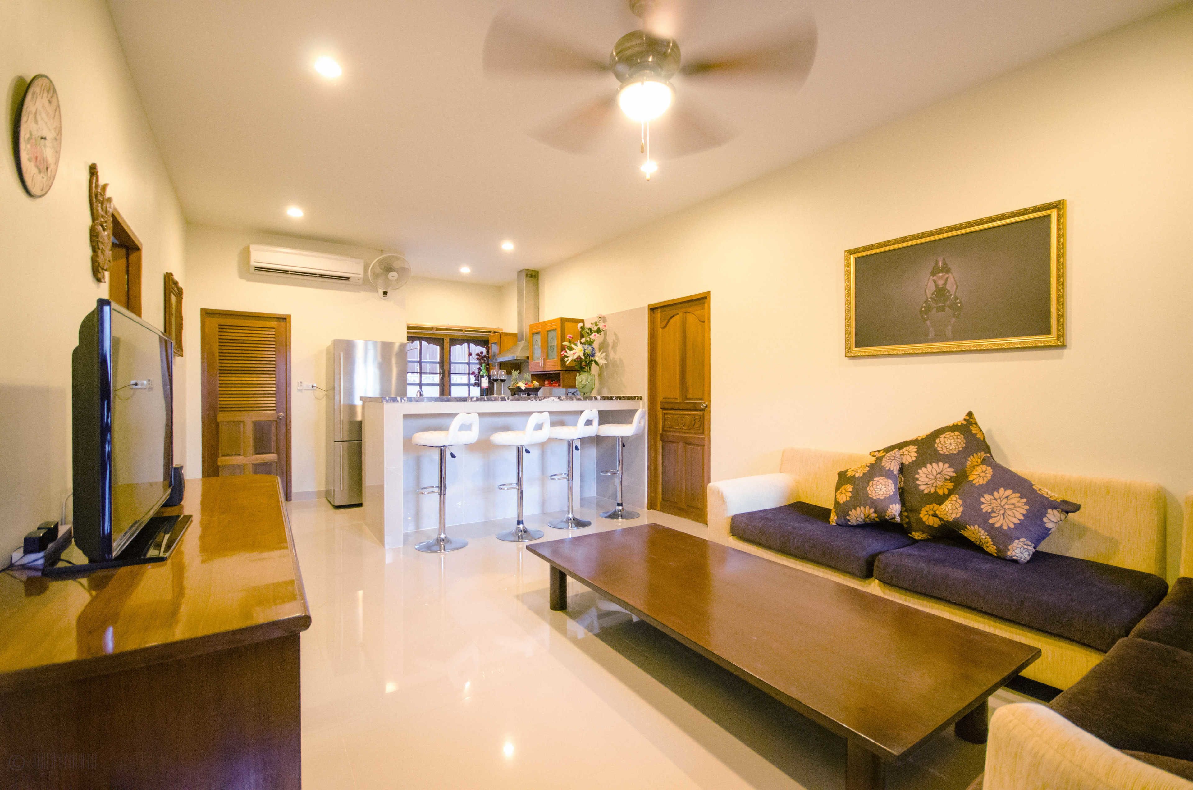 A modern living room with a sofa, a coffee table, and a TV. There is a kitchen area with a bar stool and a refrigerator.