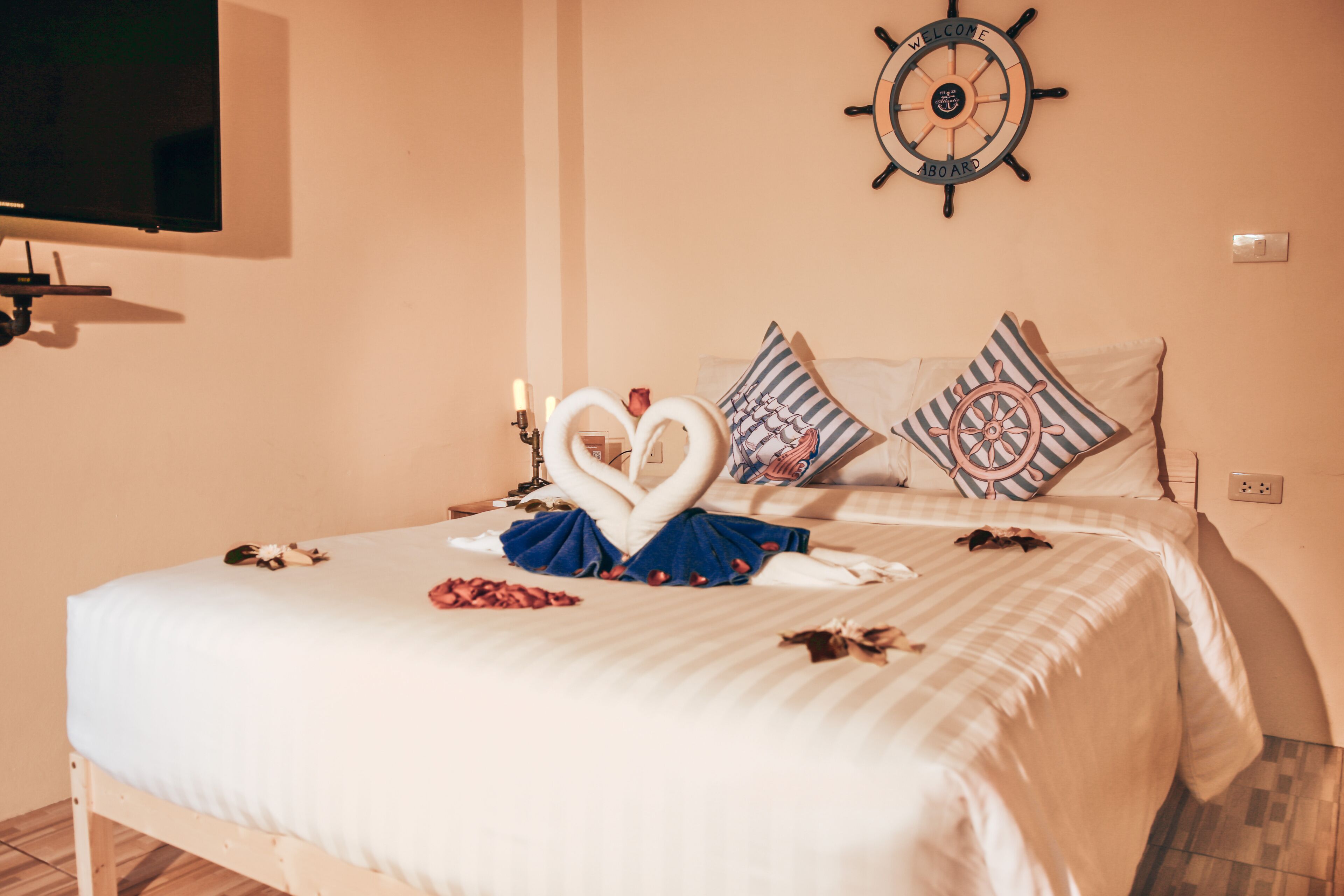 A neatly made bed with a heart-shaped towel arrangement, a ship's wheel decoration, and a television mounted on the wall.