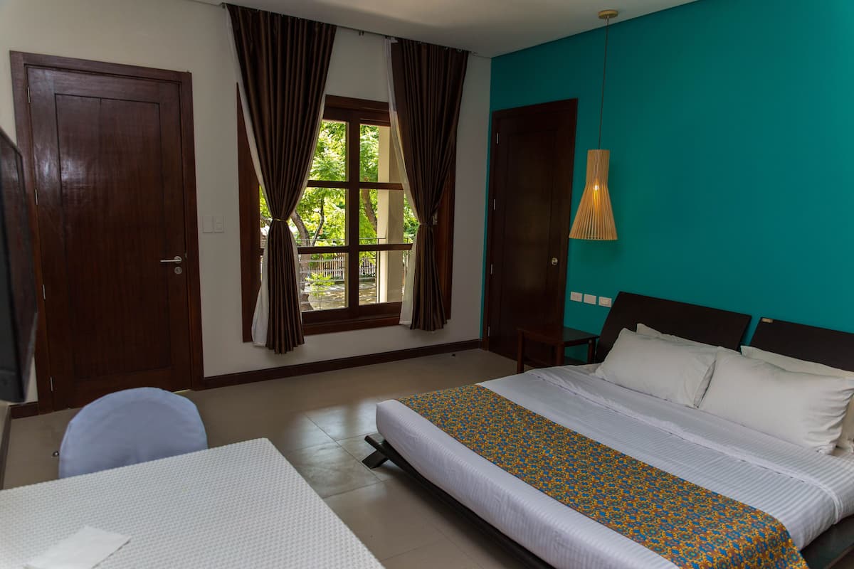 Une chambre d’hôtel avec un lit, un bureau, une chaise, une fenêtre avec des rideaux et un mur de couleur turquoise.