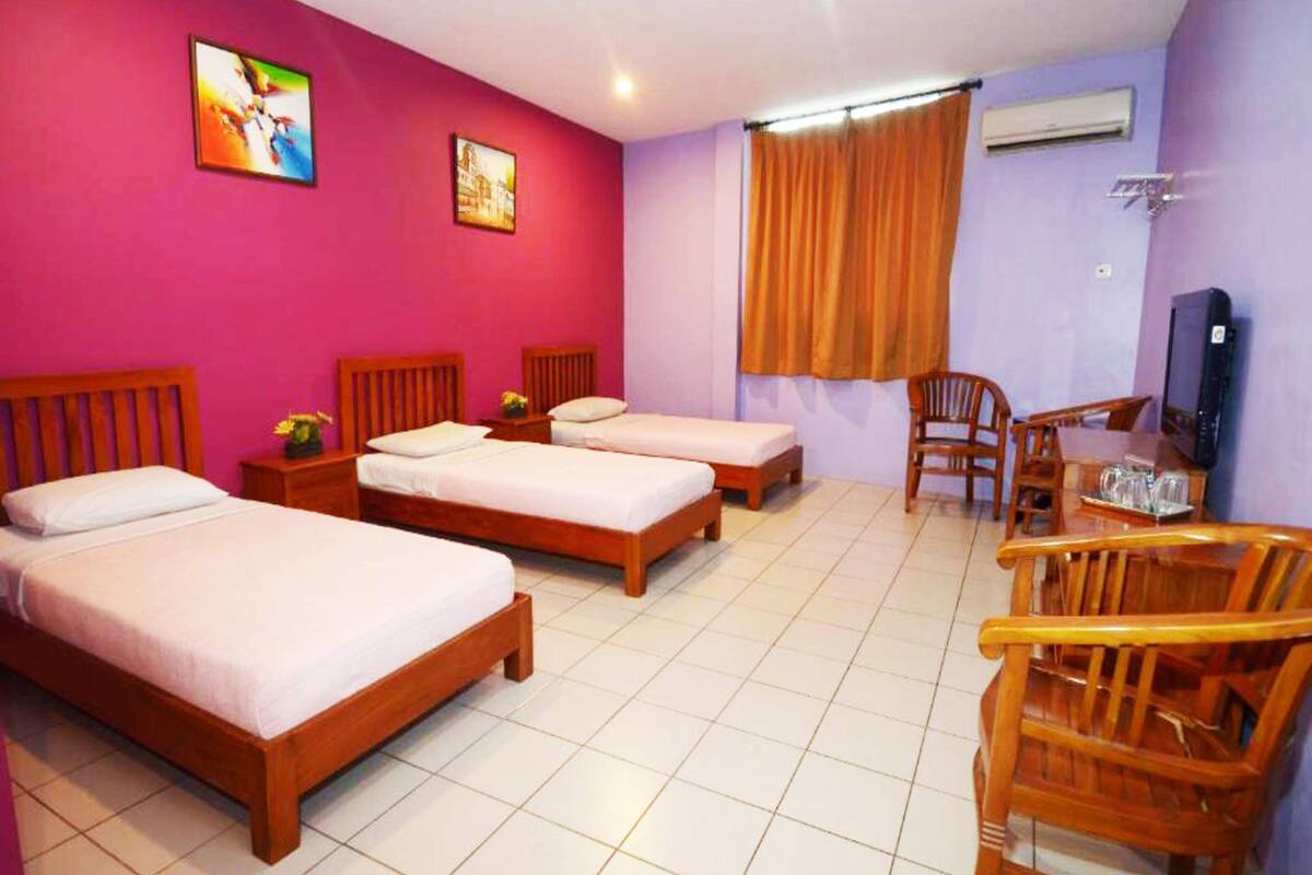 A hotel room with three wooden beds, a dining table with chairs, a television, and an air conditioning unit.