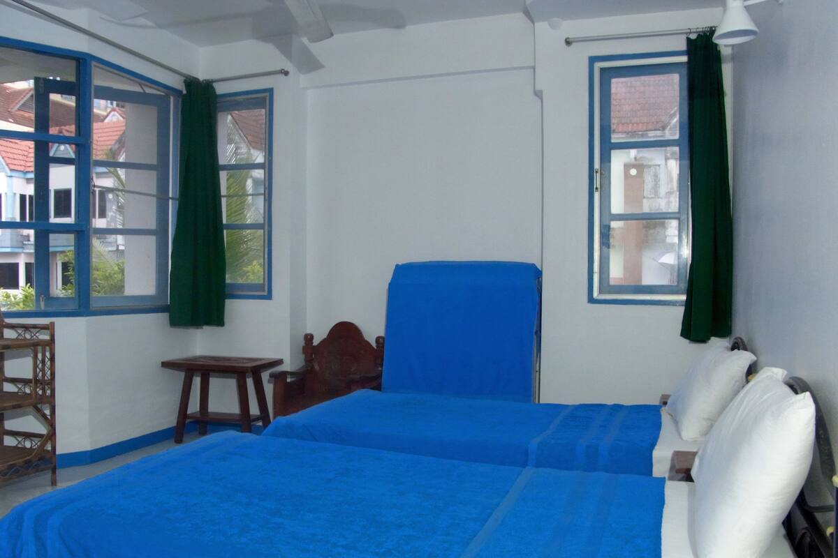 Una habitación con cama, una silla, una mesita y dos ventanas con cortinas.