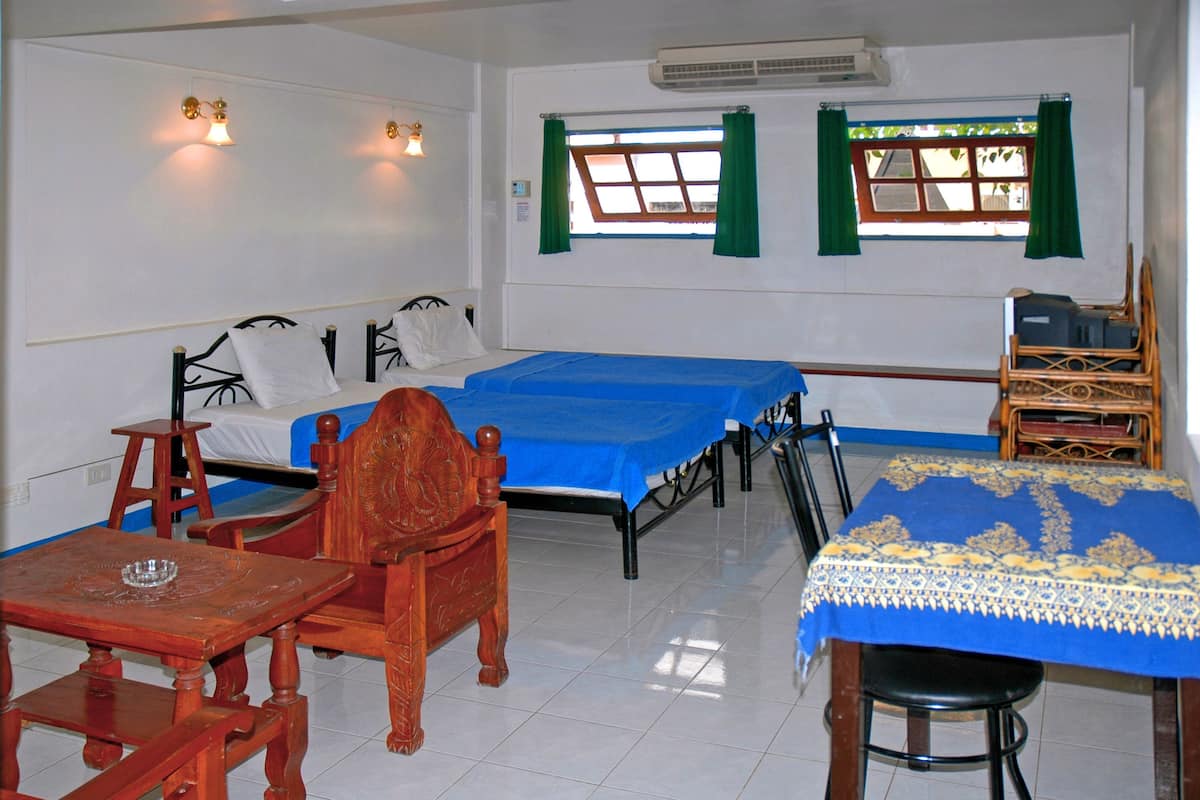 Una habitación con dos camas, una mesa de madera, una silla y un pequeño escritorio con otra silla. Hay dos ventanas con cortinas y un equipo de aire acondicionado en la pared.
