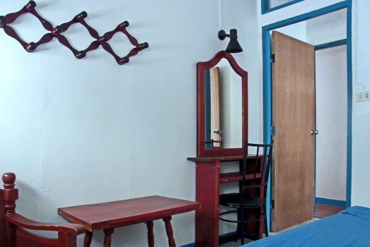 Un dormitorio con una cama de madera, una pequeña mesa de madera, una silla, un espejo y una puerta.