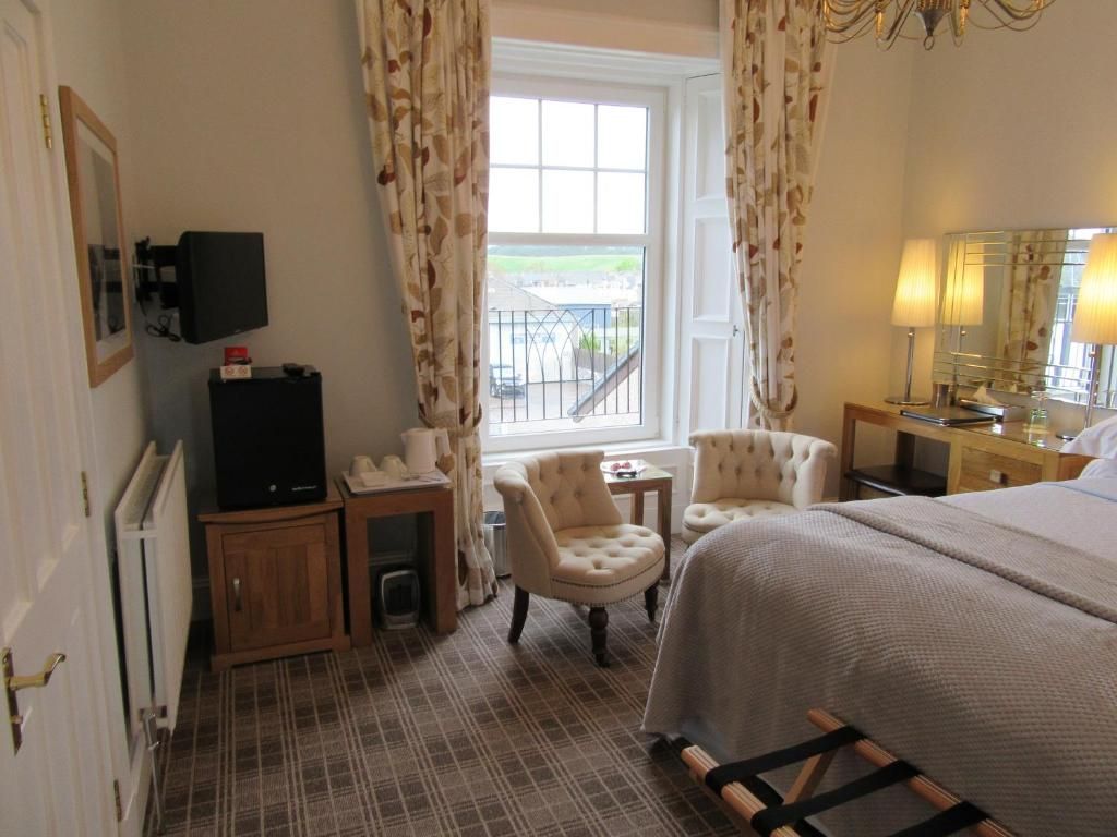 A hotel room with a bed, two armchairs, a television, a small table, a window with curtains, and a balcony view.