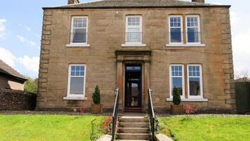 A two-story stone building with a central entrance, a staircase leading up to it, and a small garden area with a few plants.