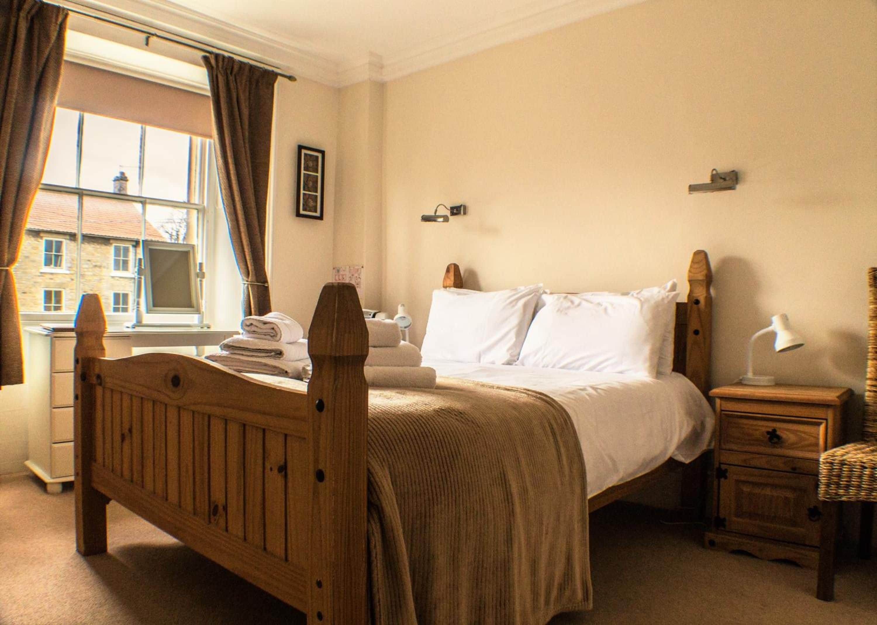 Une chambre à coucher avec un lit en bois, des tables de chevet, une lampe, une fenêtre avec des rideaux et une vue sur les bâtiments à l’extérieur.