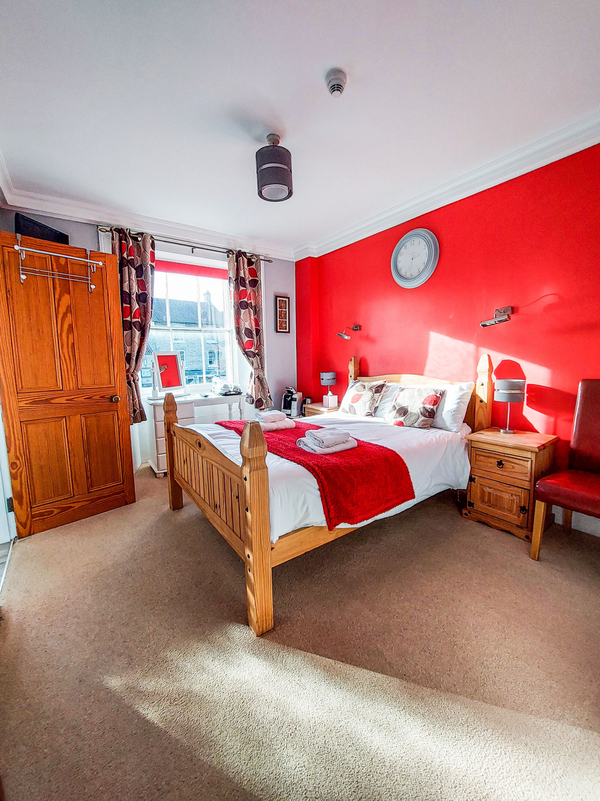 A bedroom with a wooden bed, a red wall, a clock, and a window with curtains.