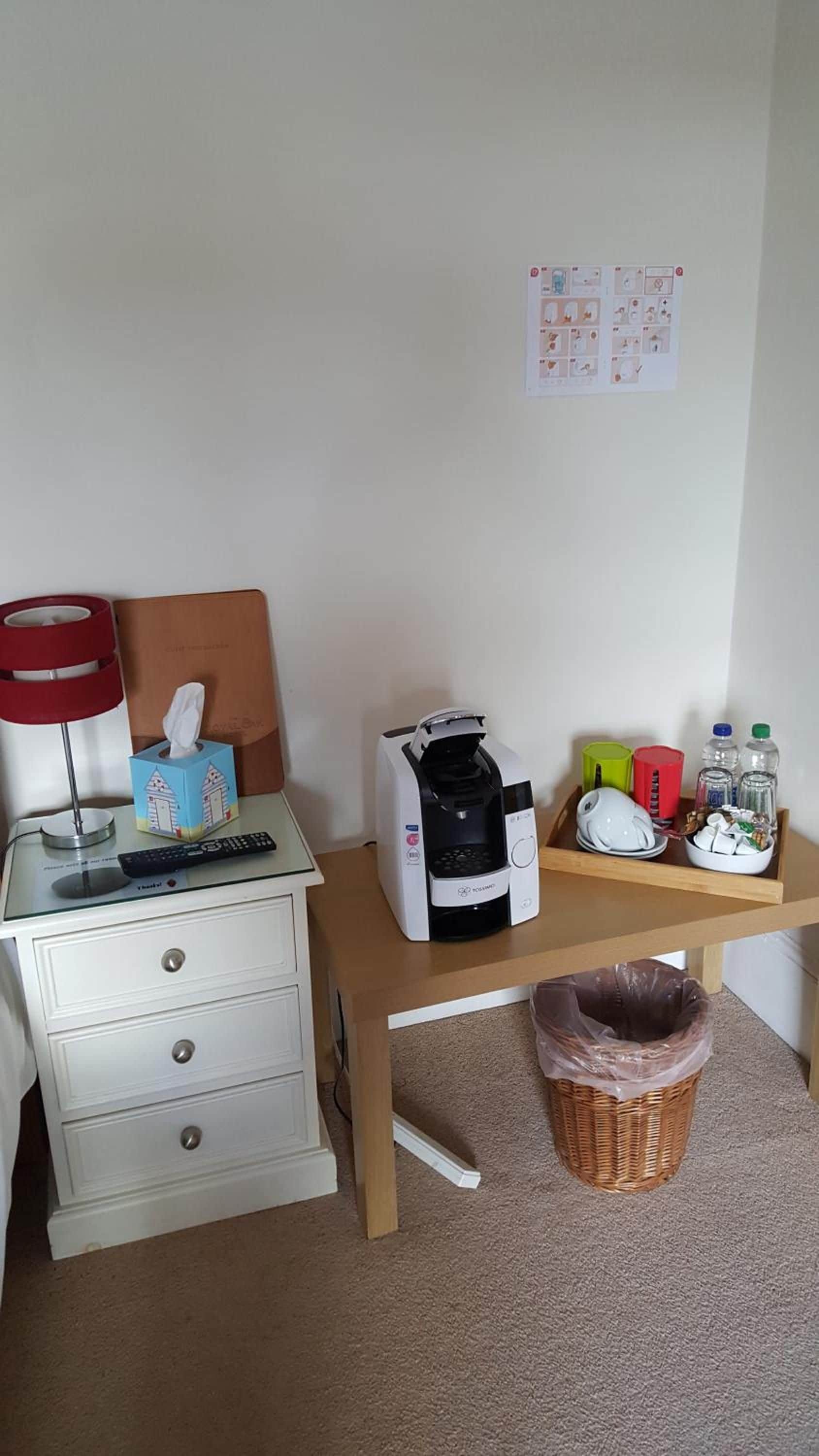 A bedroom with a bed, bedside table, a coffee maker, a tissue box, a remote control, a basket, and a wall with a notice.