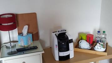 A bedroom with a bed, bedside table, a coffee maker, a tissue box, a remote control, a basket, and a wall with a notice.