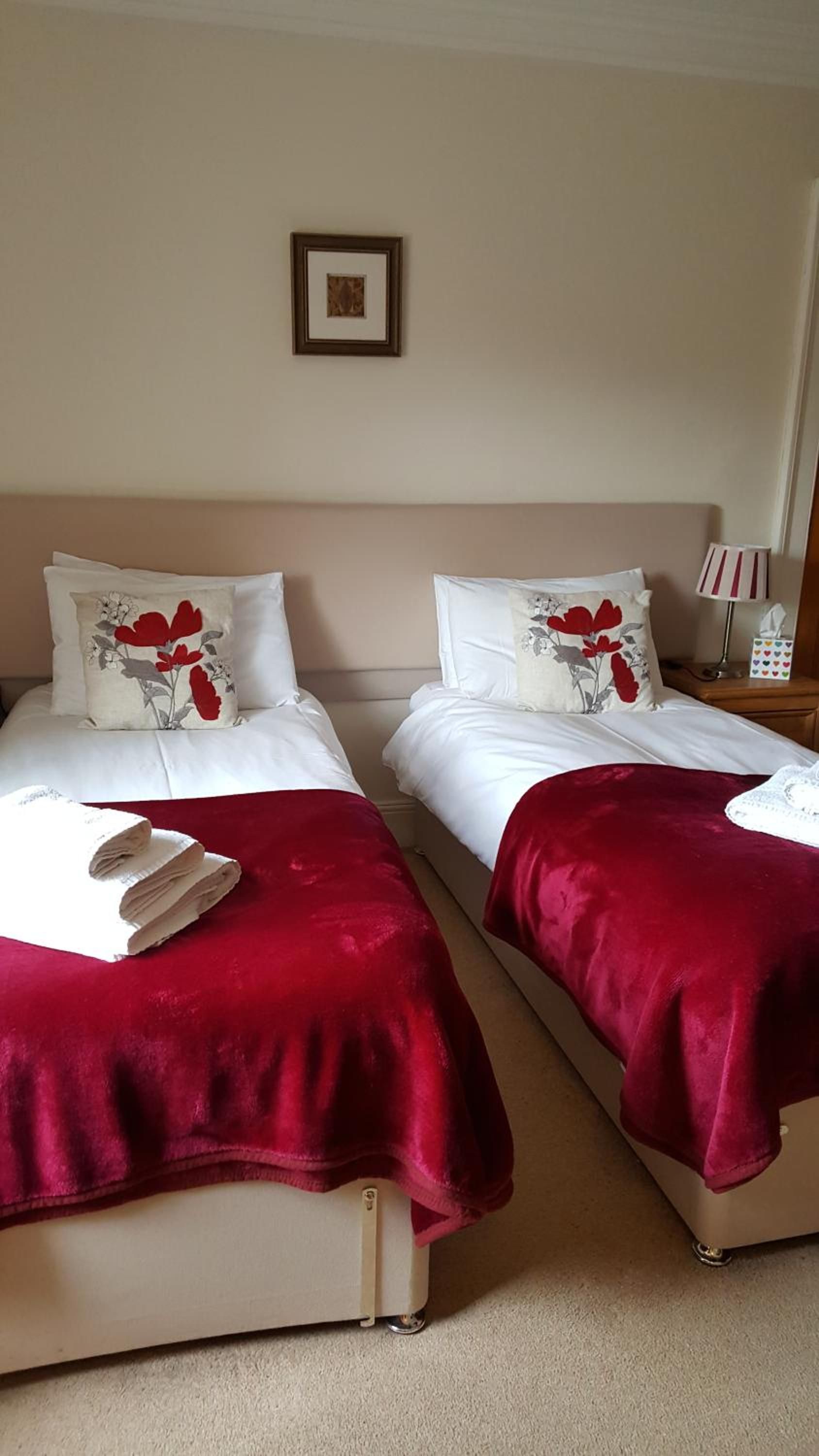 Una camera d'albergo con due letti, ciascuno con una coperta rossa e biancheria bianca. Sopra i letti, una cornice con un quadro.