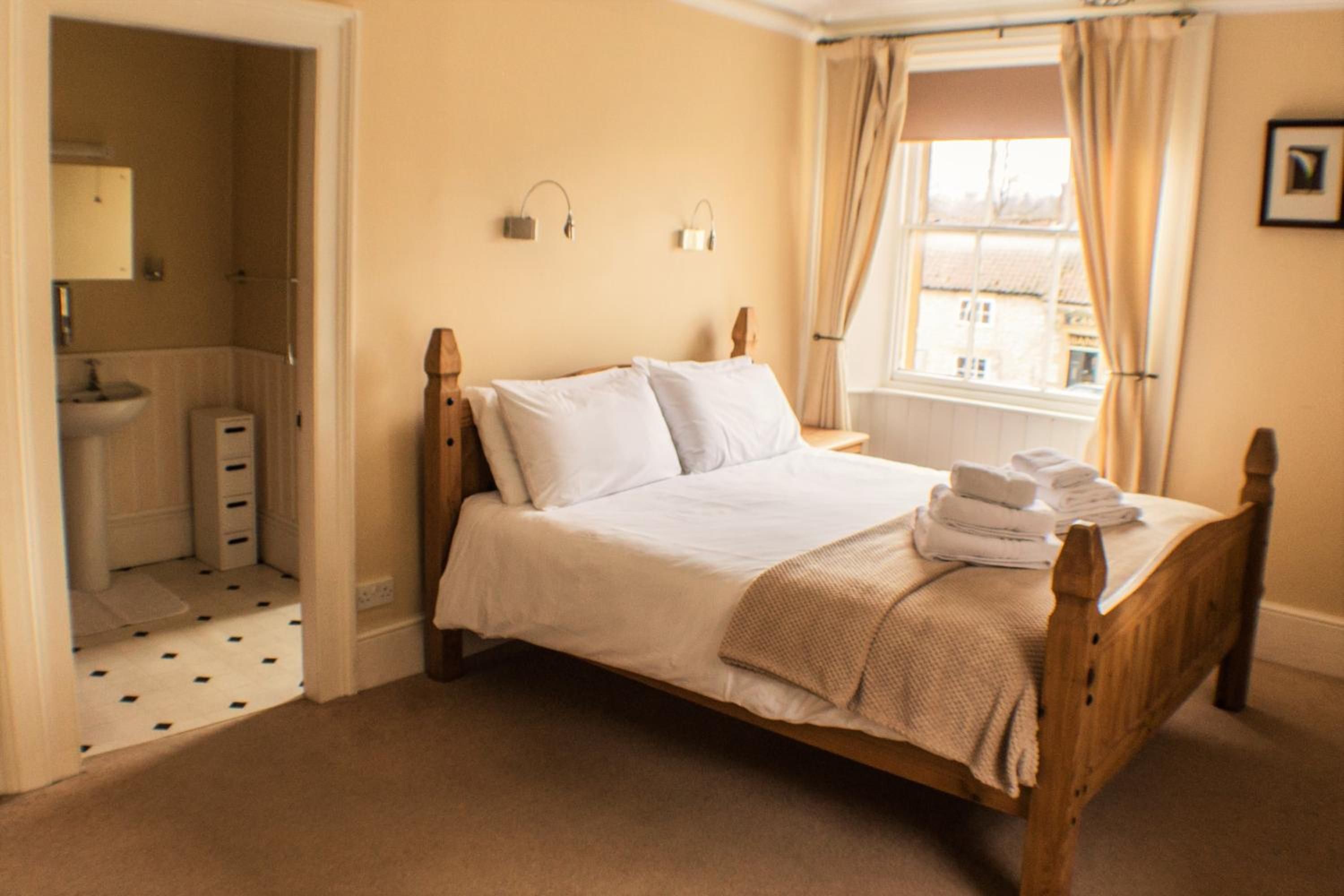 A bedroom with a wooden bed, white linens, a bathroom with a sink and mirror, and a window with curtains.