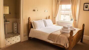 Une chambre à coucher avec un lit en bois, du linge de lit blanc, une salle de bain avec un lavabo et un miroir, et une fenêtre avec des rideaux.