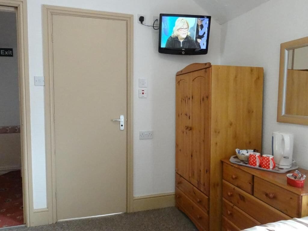 A bedroom with a wooden wardrobe, a television mounted on the wall, and a bedside table with cups and a kettle.
