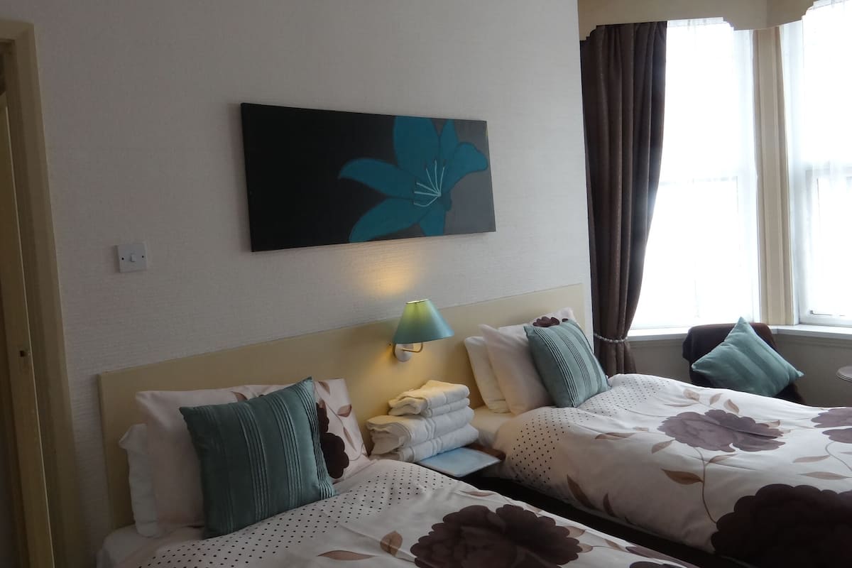 A hotel room with two beds, a wall-mounted artwork, a nightstand with a lamp, and a window with curtains.