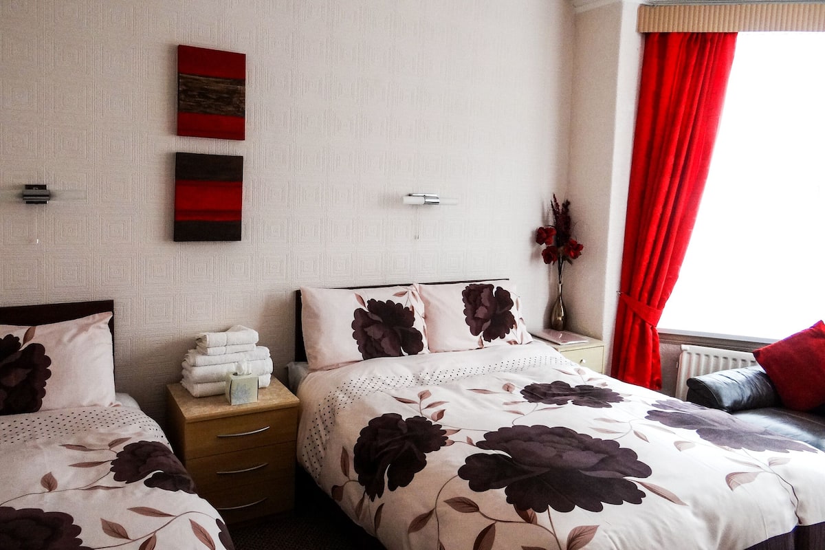 A bedroom with two beds, a nightstand, a vase with flowers, and a window with red curtains.