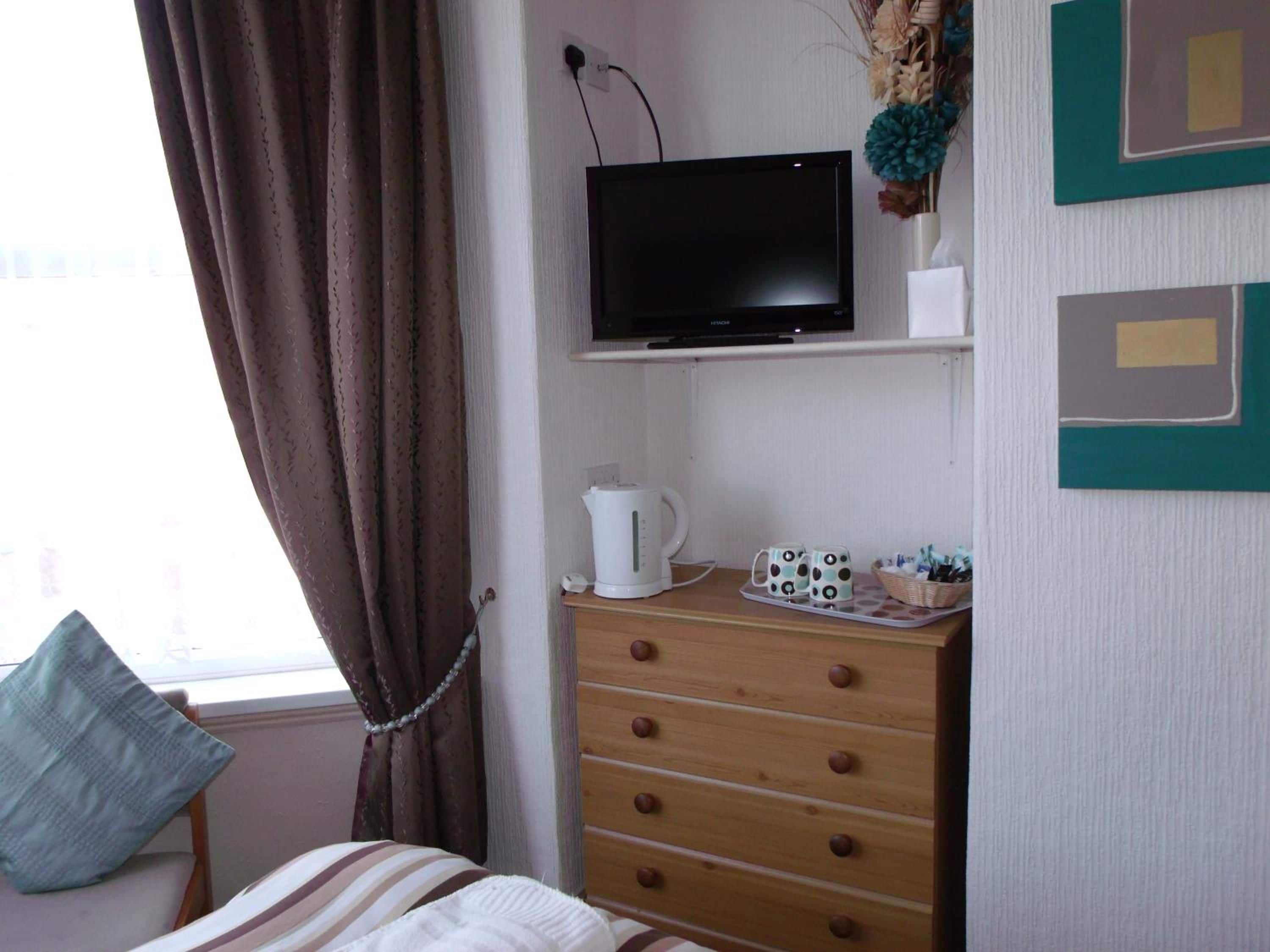 A room with a bed, a TV on a stand, a dresser with drawers, a window with curtains, and a wall with framed artwork.