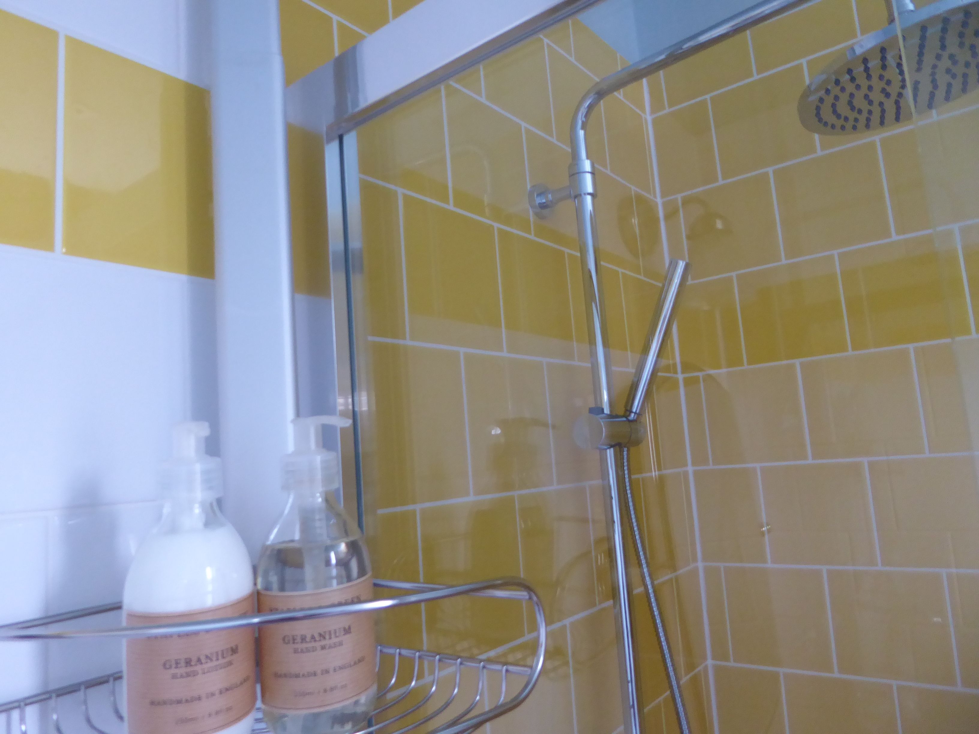 A bathroom with yellow-tiled walls, a shower with a handheld showerhead, and a shelf with two bottles of hand soap.
